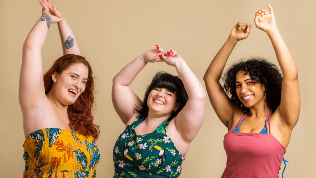 three women of varying sizes and skin tones, holding arms up cheerfully and smiling body acceptance Evoke Mind and Body Wilmington, NC